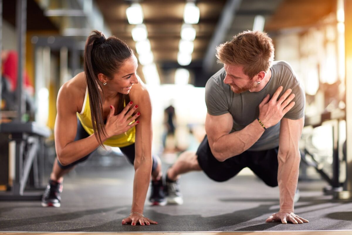 Mann und Frau trainieren gemeinsam im Fitnessstudio, lächelnd bei Plank-Übung.