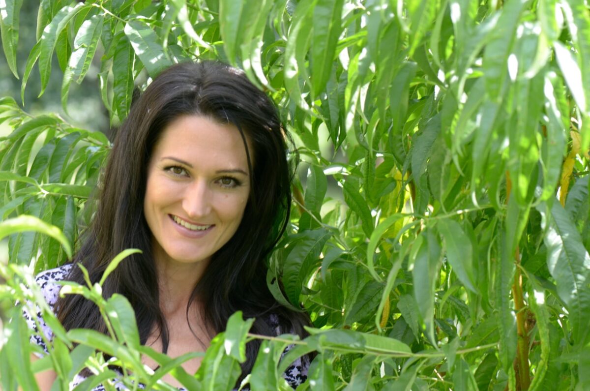 Frau mit dunklem Haar lächelt zwischen grünen Blättern hindurch.